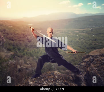 Wushu-Meister mit Schwert, Meditation auf Berggipfel, Martial Arts. Mann in schwarzem Tuch Posen mit Klinge Stockfoto