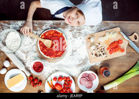 Draufsicht der junge Pizzabacken mit Pizza-Zutaten, Tomaten, Salami und Pilze auf hölzernen Tischplatte Stockfoto