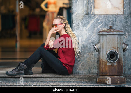 Junge Frau mit Sonnenbrille mit blonden Dreadlocks sitzt nachdenklich auf der Straße vor dem Bekleidungsgeschäft. Stockfoto