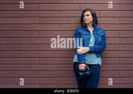 Nachdenklich Brünette kaukasische Mädchen in eine blaue Jeans-Jacke mit Digitalkamera in Händen in der Nähe von Braun strukturierte Wand. Stockfoto