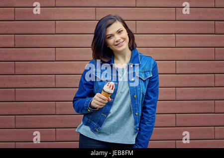 Kaukasische Brünette Mädchen hält in einer Hand leckeres Eis mit roten Früchten Marmelade in eine blaue Jeans-Jacke in der Nähe von Braun strukturierte Wand lächelnd. Stockfoto