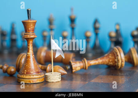 Alte hölzerne Schachfiguren auf einem alten Schachbrett. Mit die weiße Fahne der Kapitulation. Auf einem blauen Hintergrund. Stockfoto