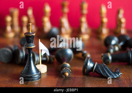 Alte hölzerne Schachfiguren auf einem alten Schachbrett. Mit die weiße Fahne der Kapitulation. Auf einem roten Hintergrund. Stockfoto