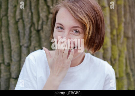 Lachende Frau in weißem Hemd mit roten Haaren über Baum. Stockfoto