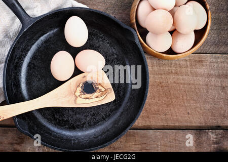 Bauernhof frische Bio braune Hühnereiern von freilaufenden Hühnern mit einer gusseisernen Pfanne und Olive Holz Spatel über einen rustikalen hölzernen Hintergrund. Stockfoto