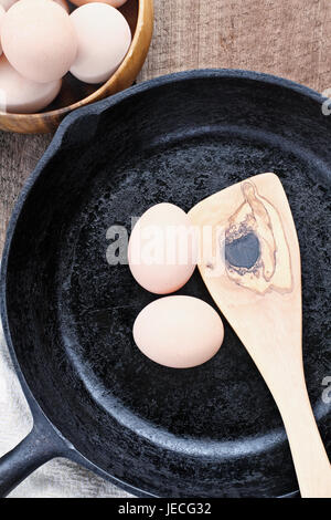 Bauernhof frische Bio braune Hühnereiern von freilaufenden Hühnern mit einer gusseisernen Pfanne und Olive Holz Spatel über einen rustikalen hölzernen Hintergrund. Stockfoto