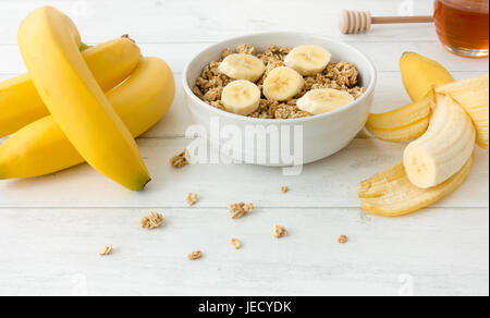 Granola Müsli mit frischen Bananen und Honig auf weißem Hintergrund aus Holz. Stockfoto