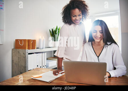 Zwei erfolgreiche Unternehmerinnen arbeiten, miteinander teilen einen Laptop-Computer in einem hellen hohen zentralen Büro lächelnd Stockfoto