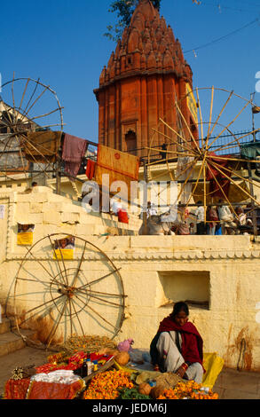 Frau verkaufen, Varanasi, Uttar Pradesh, Indien Blumen Girlanden und Weihrauch am Straßenrand stand, gelb gestrichene Gebäude und Tempel hinter. Stockfoto