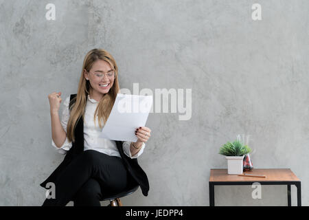 Glücklich Geschäftsfrau mit angehoben ja Geste Hand lesen Buchstabe am Schreibtisch im Büro. Unternehmerin Freude gute Nachrichten aus der Korrespondenz. Exi Stockfoto