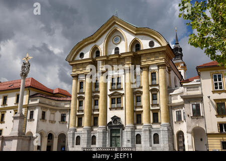 Volle Sonne auf die Ursulinen-Kirche der Heiligen Dreifaltigkeit mit Marmor-Statuen der Heiligen Dreifaltigkeit Spalte in Ljubljana Slowenien mit dunklen Wolken Stockfoto