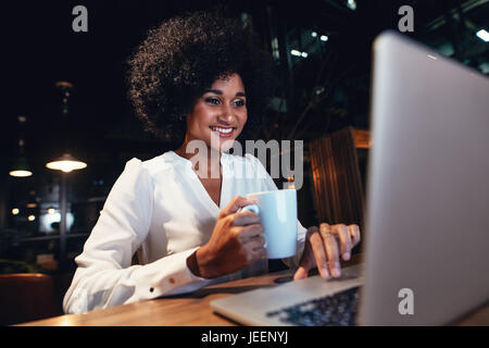 Junge Geschäftsfrau auf dem Laptop am Ende Nachtschicht arbeiten. glückliche Frau am Schreibtisch hält Kaffee und Blick auf Laptop. Stockfoto