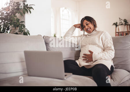 Porträt von glücklichen schwangeren Frauen auf Sofa sitzen und mit Blick auf Laptop. Frau erwartet ein Baby auf Couch sitzen und mit Laptop zu Hause. Stockfoto