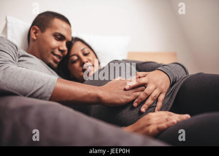 Junge schwangere paar auf Bett zu Hause mit den Händen auf dem Bauch liegend. Junges Paar ein Baby erwartet. Stockfoto