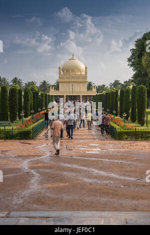 Mysore, Indien - 26. Oktober 2013: Grüne und rote Park mit Weg zu Creme gelb Tipu Sultan Mausoleum mit Kuppel unter schweren Wolkengebilde. Menschen walki Stockfoto