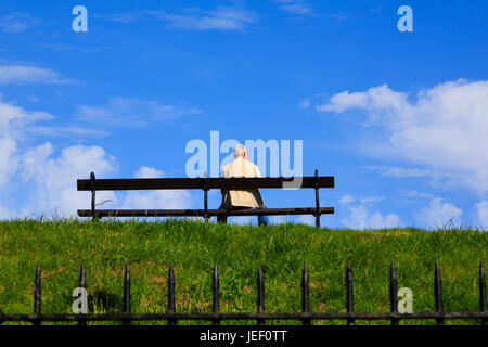 Seniorenmann, der allein auf einer Bank auf einer Grasbank vor einem blauen Himmel sitzt. An den Wänden von Berwick-upon-Tweed. Die nördlichste Stadt Englands. Stockfoto