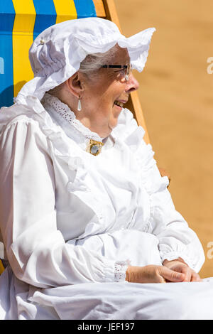 Ältere Frau, 70 Jahre, trägt Viktorianischen Dienstmädchen und Rüschen hat im Liegestuhl sitzen, Reden, von der Seite. Close-up. Teil von Broadstairs Dickens Woche. Stockfoto