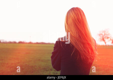 Rückansicht der eine schlanke junge Frau stehend Suche am Horizont in einem Park. Kopieren Sie Raum. Linseneffekt. Sie hat blonde oder rote Haare und trägt ein Kinderbett oder Marmelade. Stockfoto