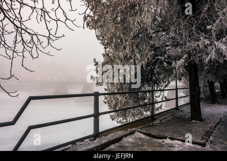 nebligen Morgen in der Nähe der Brücke über den zugefrorenen Fluss. Baum im Raureif auf dem verschneiten leer Damm. wunderschöne Stadtbild Sonnenaufgang Stockfoto