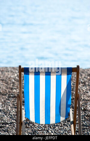 Einsam, leer, blau und weiß gestreiften Liegestuhl sitzen auf einem Kiesstrand an einem sonnigen Tag mit Meer im Weichzeichner im Hintergrund. Stockfoto