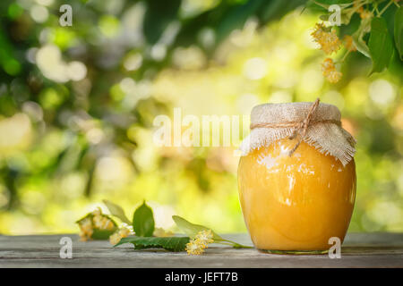 Lindenhonig. Glasdose mit Honig und Lindenblüten Blumen auf Holztisch mit unscharfen Sonnenschein blühende Linde auf dem Hintergrund. Foto mit Kopie spac Stockfoto