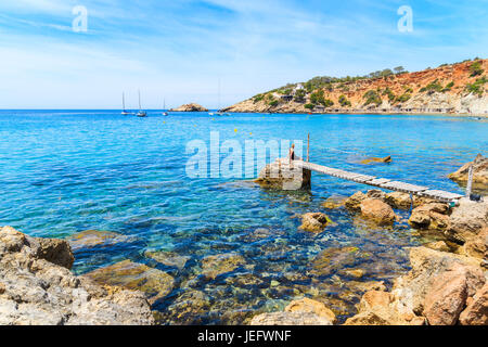 Unbekannte Frau sitzen auf kleinen Holzmole in Cala d ' Hort Bucht und Blick auf Es Vedra Insel Ibiza, Spanien Stockfoto