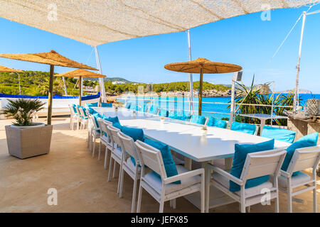 Strand von CALA NOVA, Insel IBIZA - 20. Mai 2017: Weißen Tisch und Stühle mit blauen Kissen auf Restaurant-Terrasse am Strand von Cala Nova mit schönen Blick aufs Meer vie Stockfoto