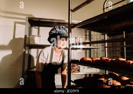 Baker steht in der Nähe von Regalen mit frischen Brötchen, lächelnd Baker entfernen Backen Tablett von Rack Stockfoto