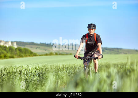 Attraktive Radfahrer fährt auf der Straße in einem Feld an einem sonnigen Tag gegen blauen Himmel. Stockfoto