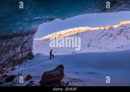 A man and the Peaks of surrounding Mountains at Sunrise werden von dem Eingang zu Einer großen Eishöhle im Augustana-Gletscher in der Alaska Range eingerahmt... Stockfoto