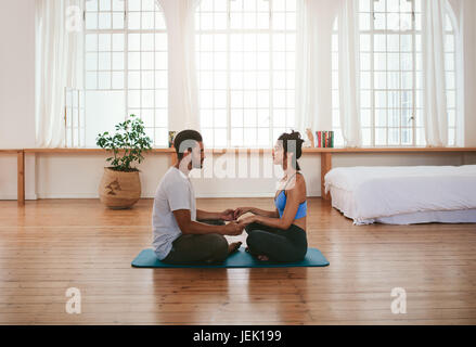 Seitlicher Blick auf schöne junge Paar Yoga zu Hause zu tun. Sie sitzen zusammen auf der Yoga-Matte, Hand in Hand. Stockfoto