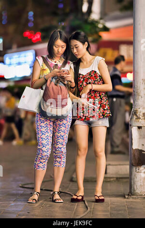 KUNMING - 10. JULI 2014. Zwei trendige Mädchen, die mit Smartphone in der Innenstadt beschäftigt sind. Stockfoto