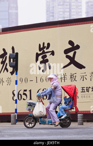 NANJING - 25. MAI 2014. Frau mit Kind auf E-Bike vor Werbewand. Außenwerbung wurde Chinas drittgrößtes Medium. Stockfoto