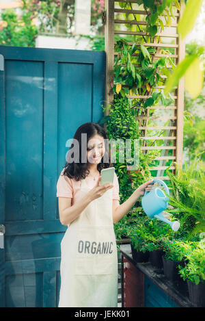 Junge asiatische Frau Gärtner mit Handy während der Bewässerung der Pflanzen Stockfoto