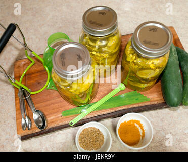 Gurken in Gläsern sitzen auf einem Schneidebrett auf einem Küchentisch zusammen mit Gurken, Geschirr und Gewürze Stockfoto