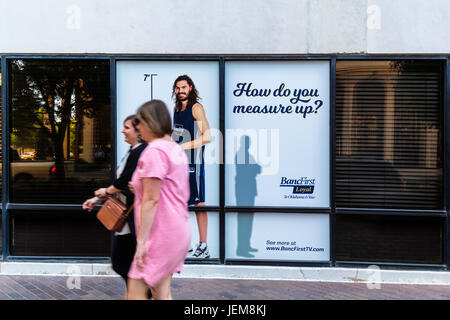 Zwei Frauen gehen vorbei an einem lebensgroßen Poster von NBA-Spieler Steven Adams im Fenster des BancFirst in der Innenstadt von Oklahoma City, Oklahoma, USA. Stockfoto