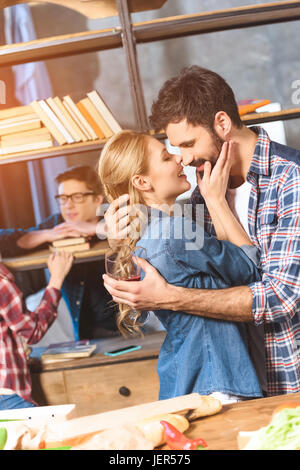 Jugendliche feiern zu Hause. Liebende Paar küssen, während andere in der Nähe von Bücherregal im Gespräch Stockfoto