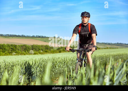Attraktive Radfahrer fährt auf der Straße in einem Feld an einem sonnigen Tag gegen blauen Himmel. Stockfoto