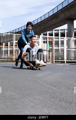 Junge Paare, die Spaß in der Stadt. Die Frau treibt ihr Partner entlang, als er auf einem Skateboard sitzt. Sie lachen beide. Stockfoto