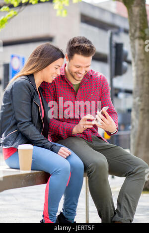 Junges Paar sitzt in der Stadt. Der Mann hält eine Smartphone, die sie beide betrachten. Stockfoto