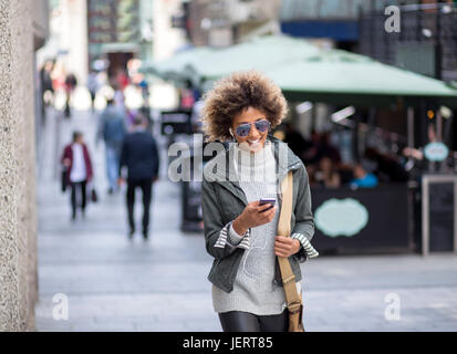 Junge Frau zu Fuß durch die High Street. Trägt sie Kopfhörer, die mit dem Smartphone verbunden sind, die sie verwenden, ist. Stockfoto