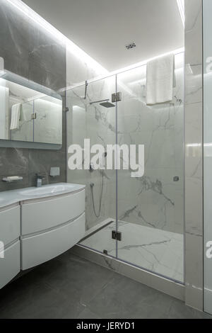 Hellen Badezimmer mit strukturierten gefliesten Wänden. Es gibt eine Dusche mit einem Glas Tür, weißen Waschbecken mit einem Wasserhahn, Spiegel, leuchtenden Lampen, Handtuch aufhängen. Indoor Stockfoto