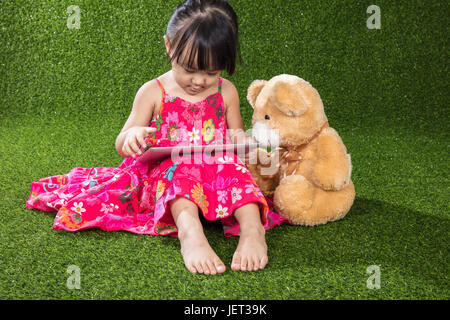 Asiatische chinesische Mädchen sitzen auf dem Rasen und Tablet-Computer mit Teddybär bei Outdoor-Park zu spielen Stockfoto