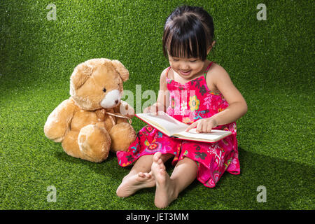 Asiatische chinesische Mädchen sitzen auf dem Rasen und Buch mit Teddybär im Outdoor-park Stockfoto
