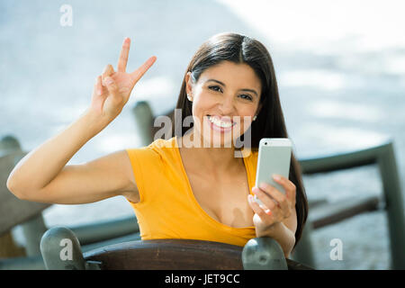 Glückliche junge Latin-Frau sprechen und SMS auf smart Phone geben das Peace-Zeichen, lässige Kleidung, sitzen mit einen nehmen Weg Kaffee in einem städtischen Stockfoto