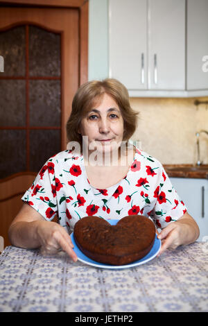 Kaukasische ältere Frau mit Herz geformten Kuchen auf der Platte während der Sitzung in häusliche Küche Stockfoto