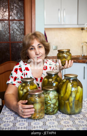 Attraktive ältere Frau zeigt Glas mit marinierten Gurken beim Sitzen am Tisch in der Küche Stockfoto