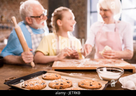 Großmutter, Großvater und Enkelin Kochen und Kneten von Teig für Kekse am Küchentisch und gebackene Plätzchen im Fach im Vordergrund, die Küche von Stockfoto