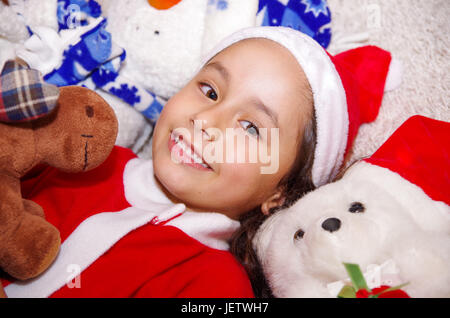 Schönes Lächeln auf den Lippen Litle Mädchen ein Weihnachten Kleidung zu tragen, einen Elch und Bär Teddy umarmen, Verlegung auf einer weißen Decke. Stockfoto