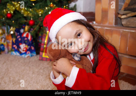 Schönes Lächeln auf den Lippen Litle Mädchen Kleidung ein Weihnachten, umarmt einen Elch Teddy mit einem Weihnachtsbaum-Hintergrund mit einigen Geschenken. Stockfoto